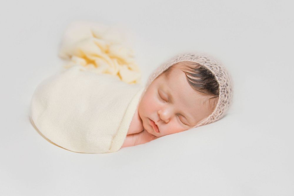 Peaceful newborn baby wrapped in cream blanket wearing knit bonnet during minimalist portrait session.