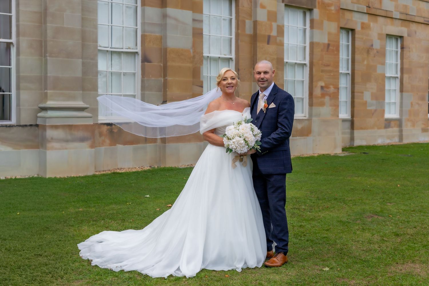 A couple in formal wedding attire poses together on a grassy lawn in front of a stone building.