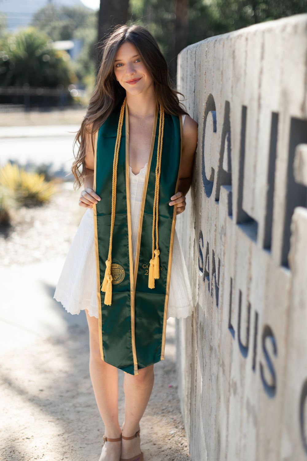 Young woman in green and gold graduation stole posing by Cal Poly San Luis Obispo sign in white dress.