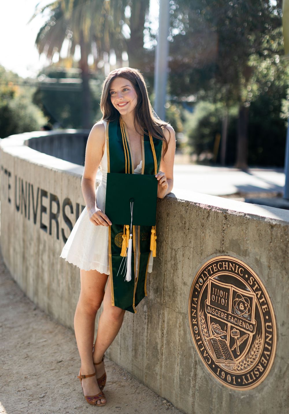Cal Poly SLO graduate poses in green and gold stole beside university seal wall outdoors.