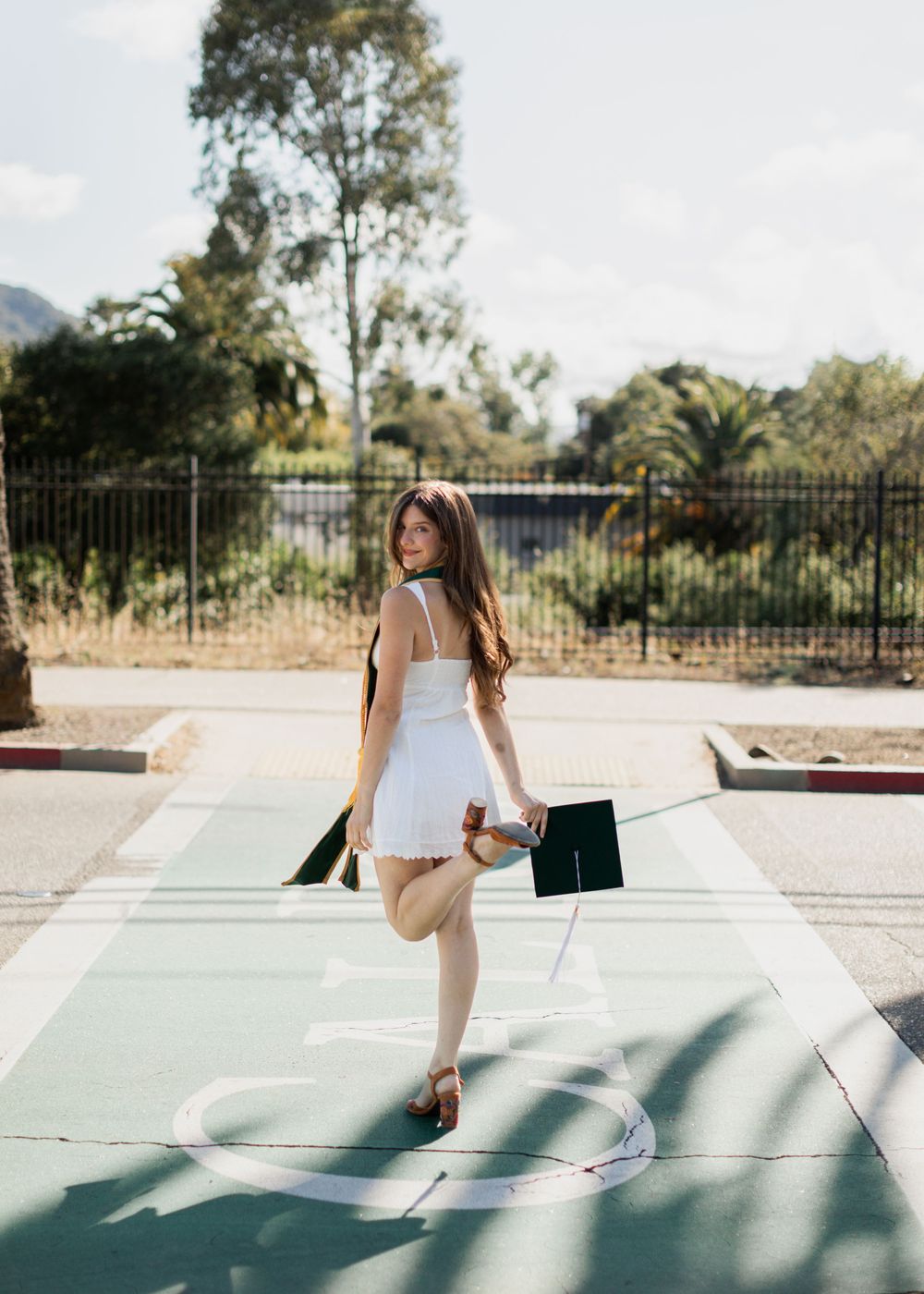 Young woman in white dress holding graduation cap poses on outdoor court at Cal Poly San Luis Obispo.
