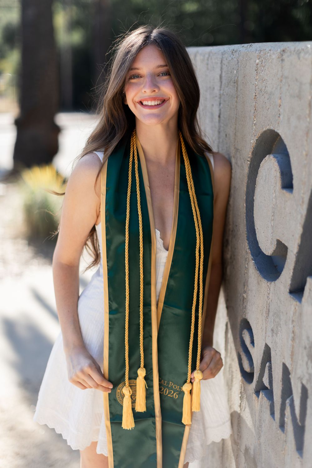Smiling young woman in white dress wearing green and gold graduation stole poses outdoors near a stone monument.