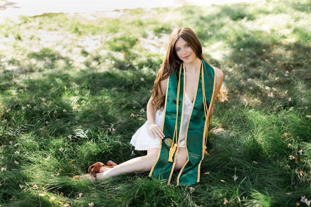 Young woman in white dress and green graduation stole sitting in lush grass for senior portrait.