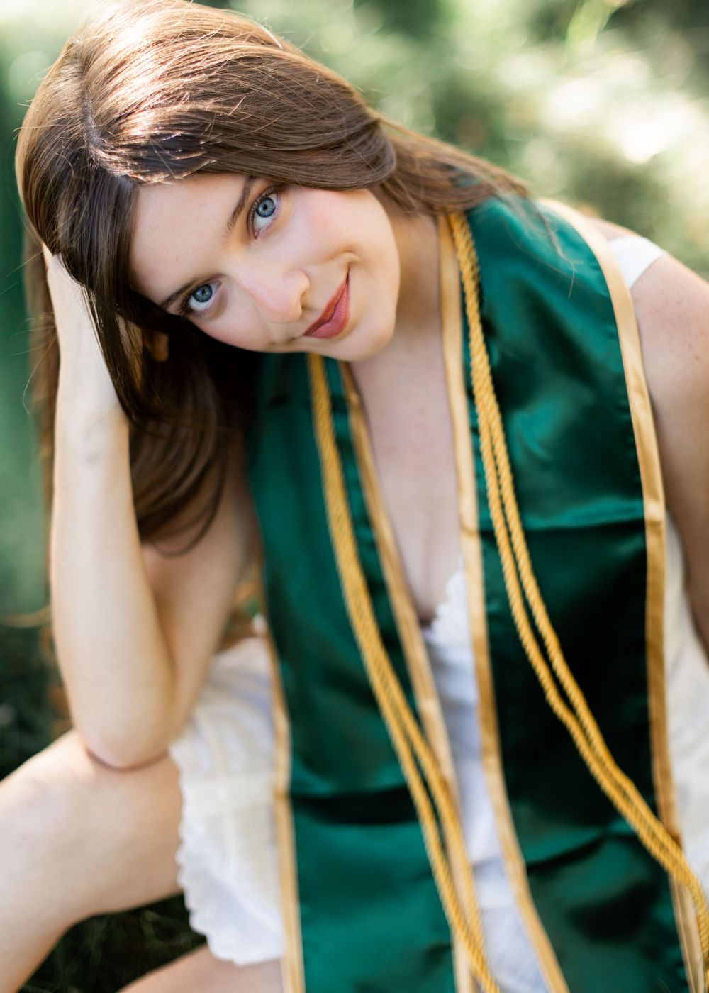 Close-up portrait of blue-eyed brunette woman wearing green and gold graduation stole outdoors.