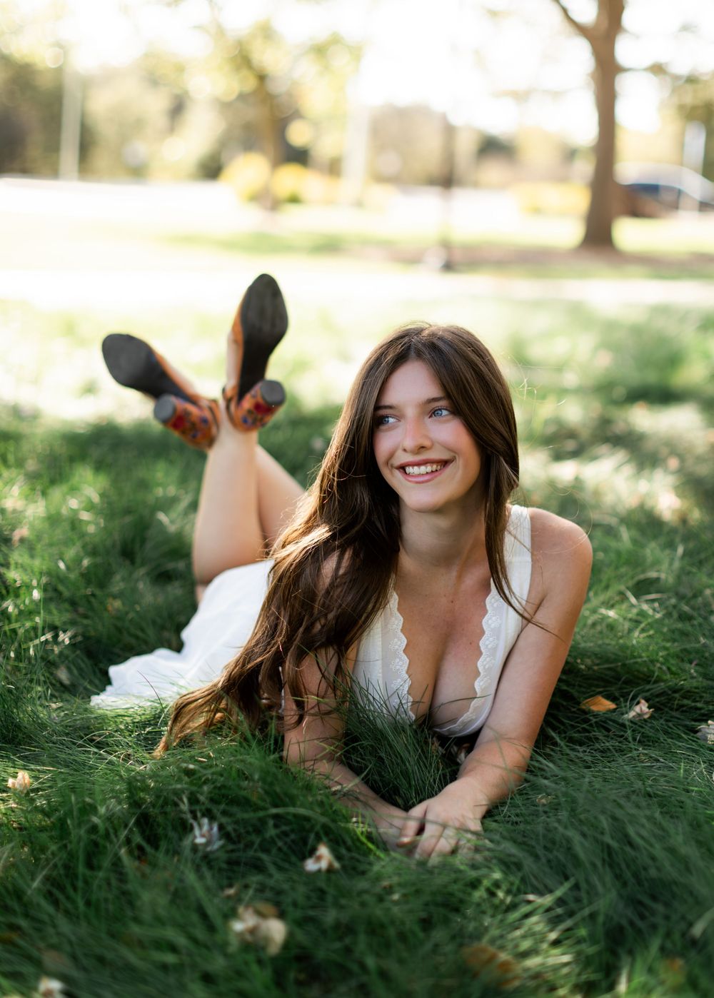 Young woman in white dress lying on green grass smiling, long brown hair, casual outdoor portrait.