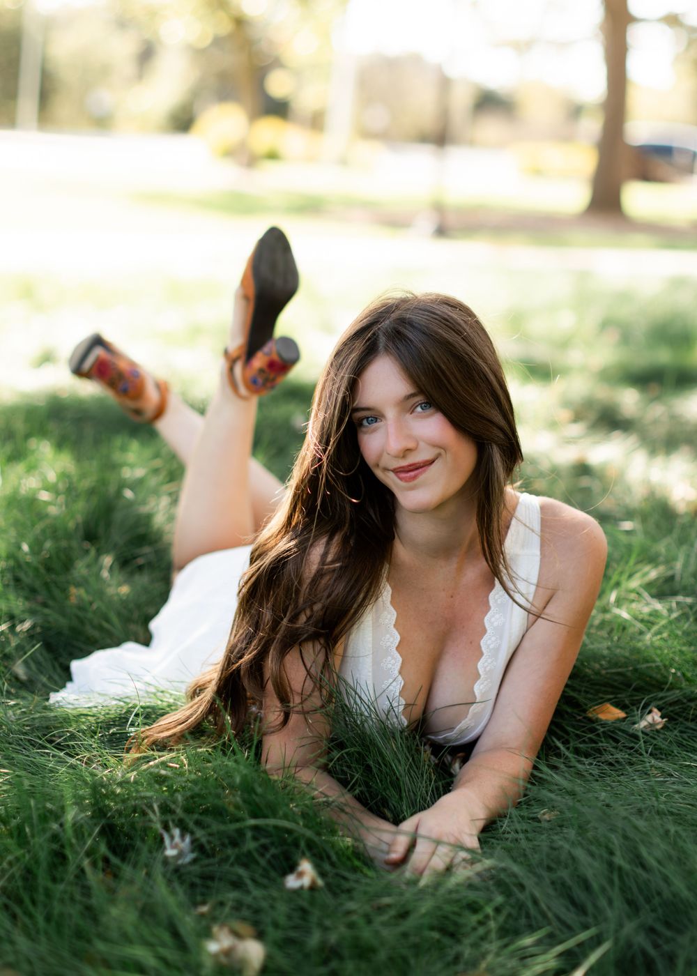 Smiling young woman in white dress lying on green grass in sunny park setting.