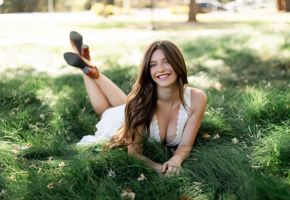 Young woman in white dress lying on lush green grass, smiling cheerfully in a sunny park setting.