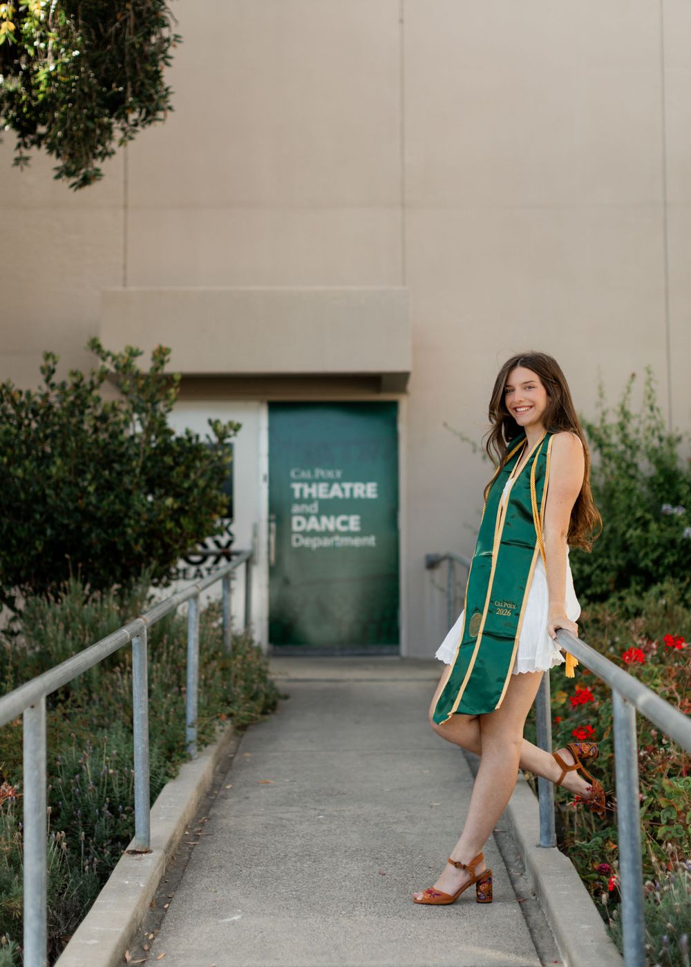 Female graduate in green stole smiles on steps outside Cal Poly Theatre and Dance Department.