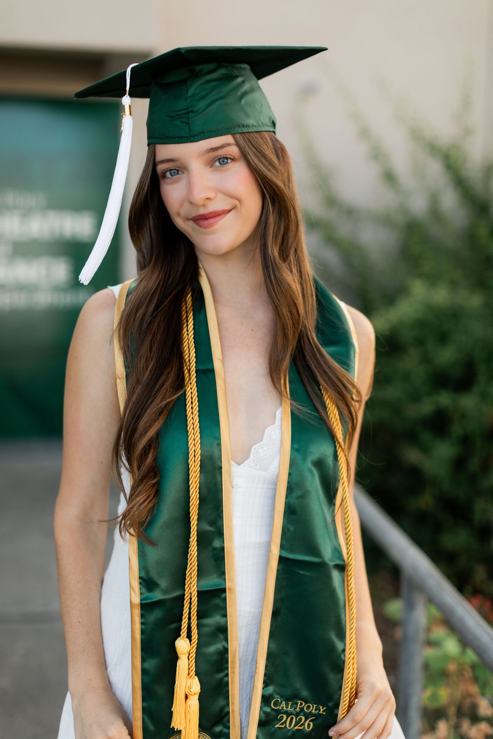 Young woman in green Cal Poly 2026 graduation cap and gown smiling outdoors.