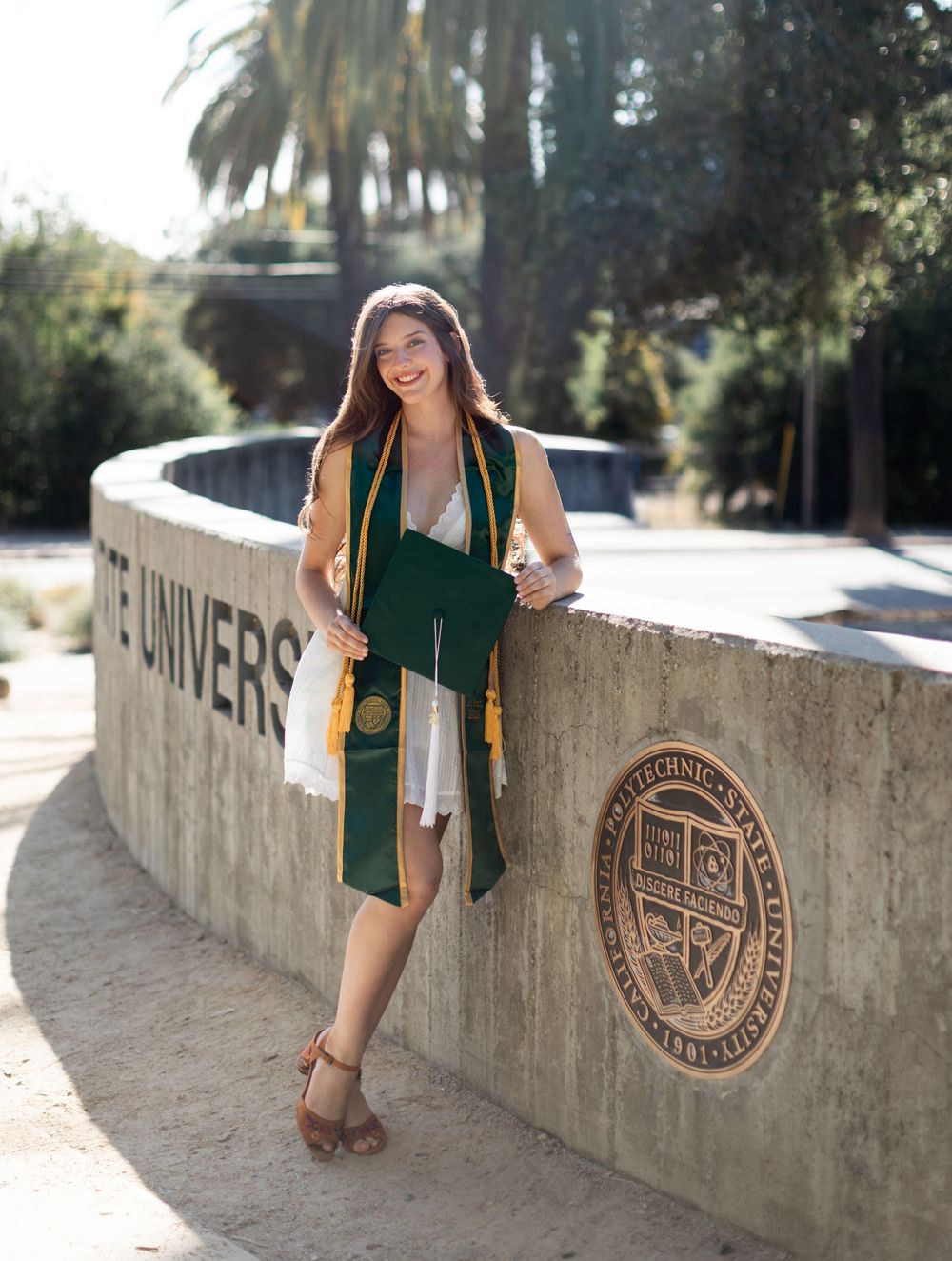 A smiling college graduate poses in green and gold regalia holding a diploma at Cal Poly State University.