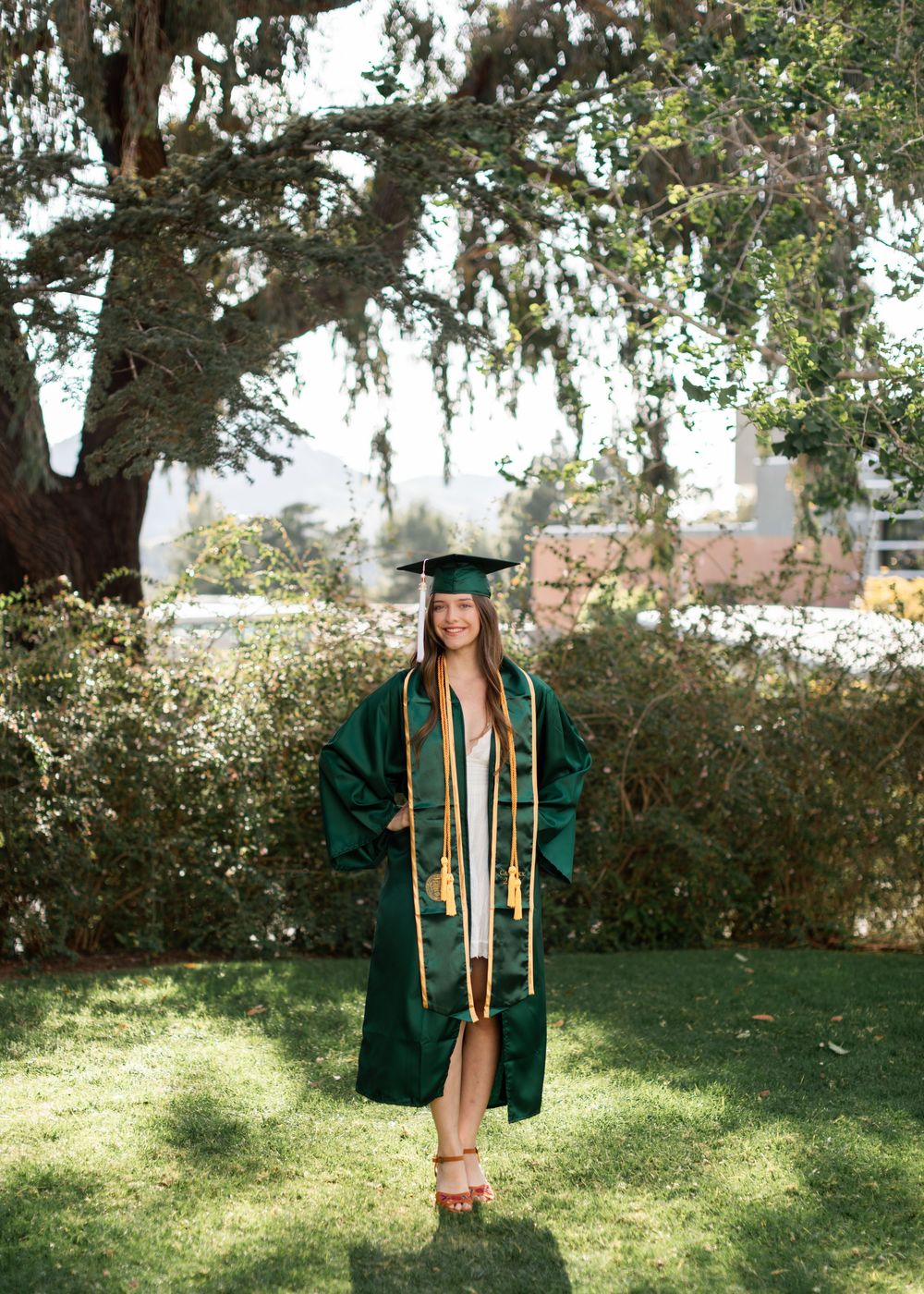 Young woman in green Cal Poly graduation cap and gown standing on grass under a large oak tree.