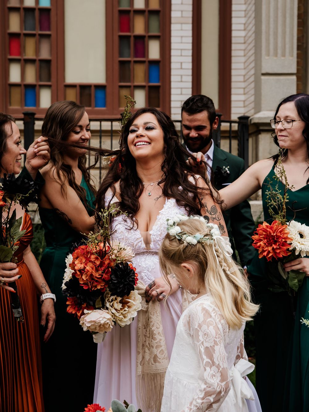 Wedding party celebrating with laughter and colorful bouquets against a brick building backdrop.