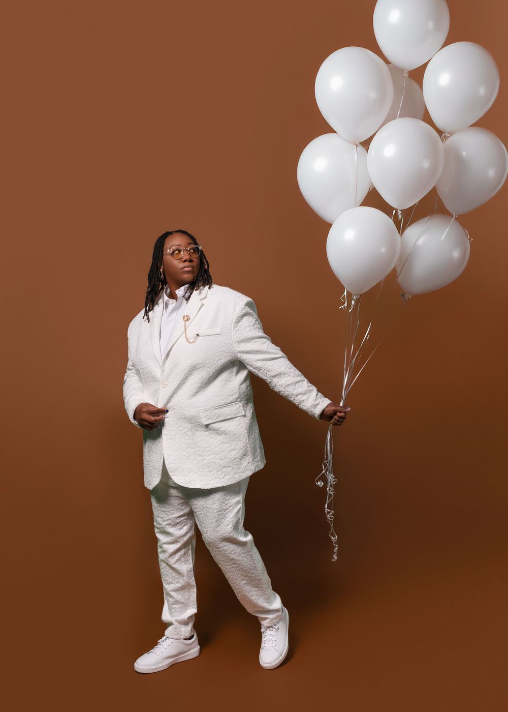Stylish figure in white suit holding white balloons against brown backdrop.