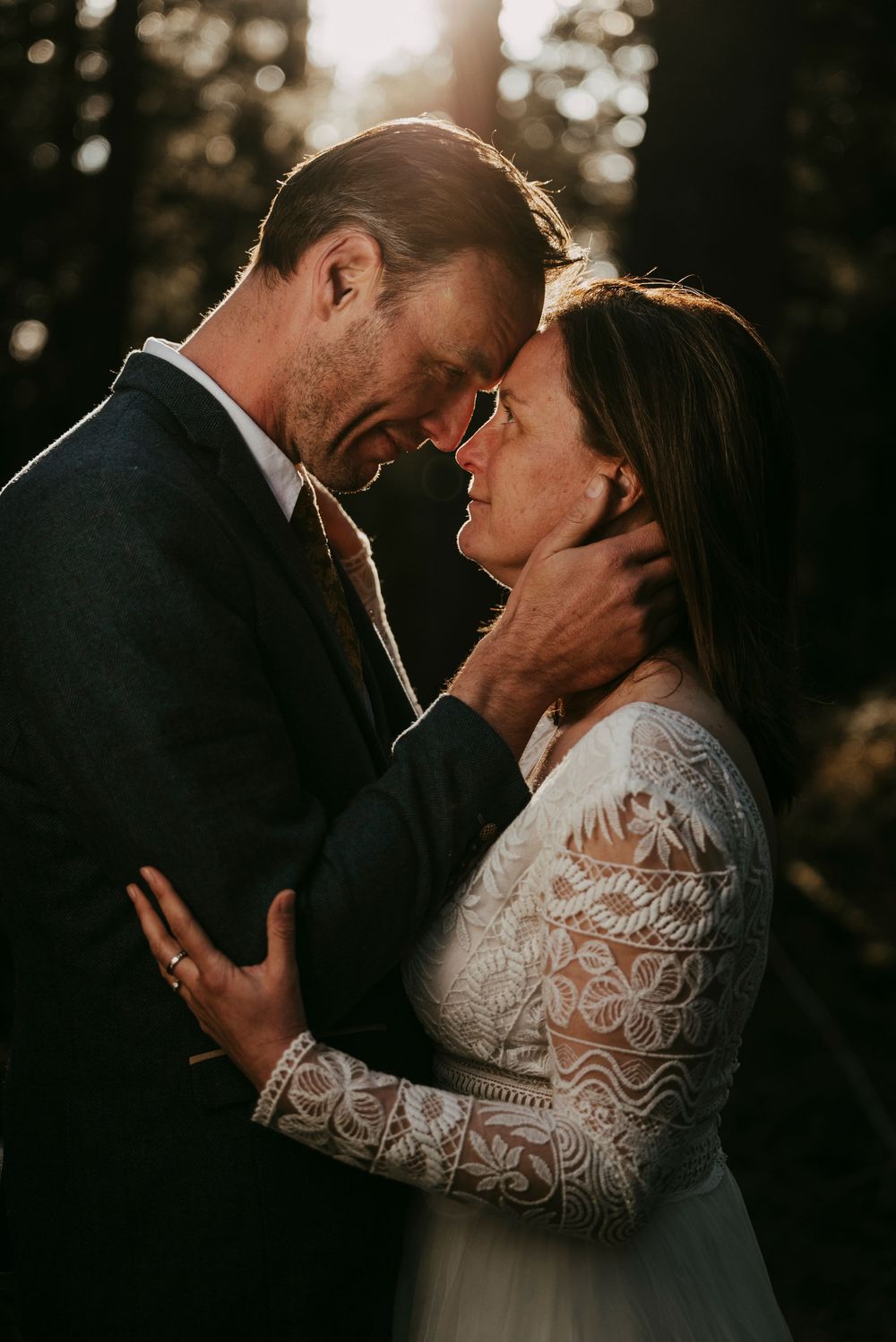 Bride and groom sharing intimate forehead touch in forest, golden backlight, lace wedding dress.