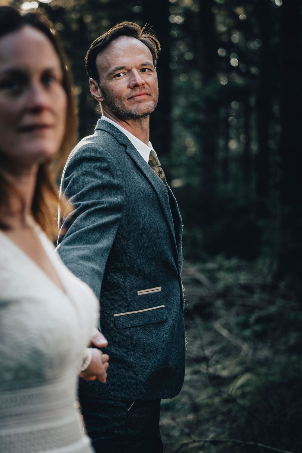 Groom in grey suit looking back at camera in forest, bride in white lace dress in foreground.