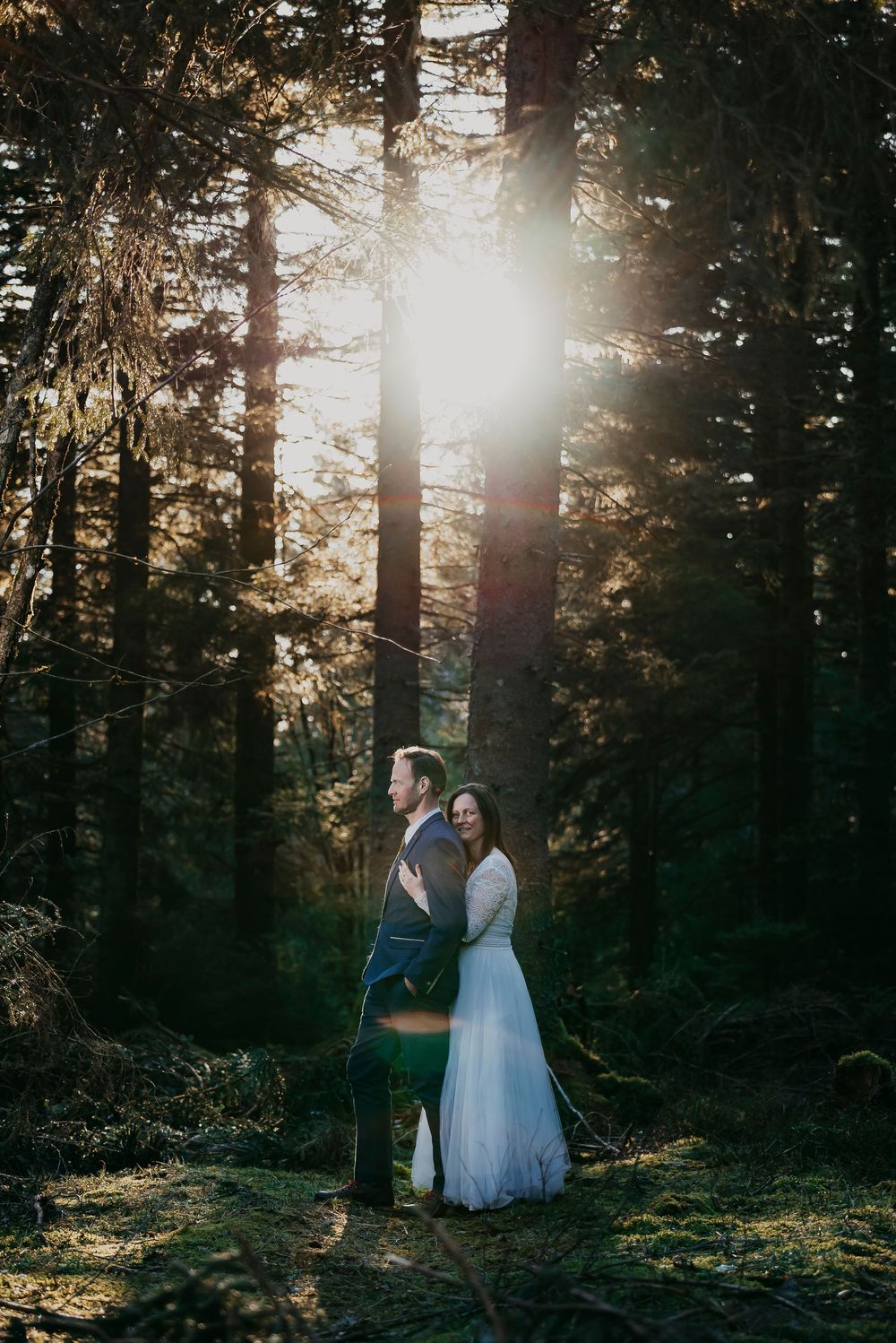 Wedding couple standing back-to-back in sunlit forest, bride in flowing white dress, groom in suit.