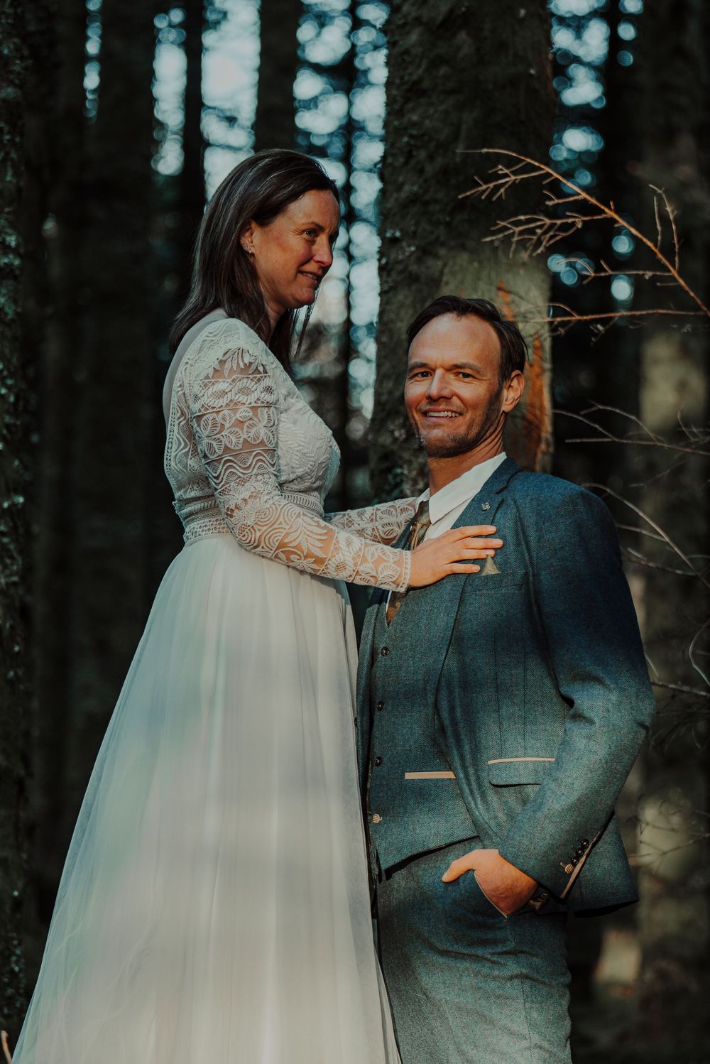 Bride in lace wedding dress smiling at groom in blue suit leaning against tree in forest.
