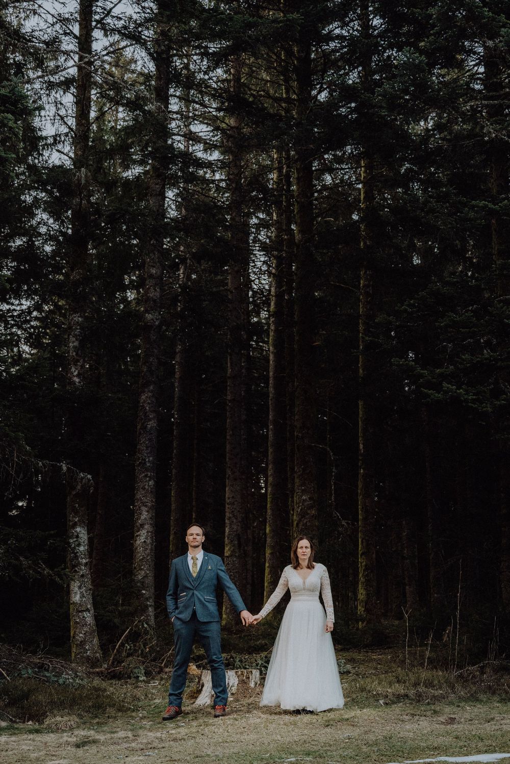 Wedding couple holding hands standing before tall pine forest, bride in white gown, groom in suit.