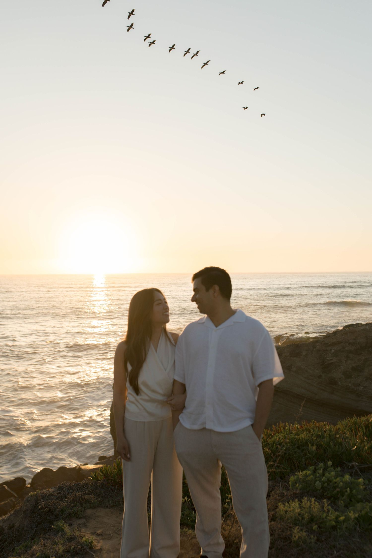 A couple in white outfits stands on a coastal cliff at sunset with birds flying overhead in a V formation.