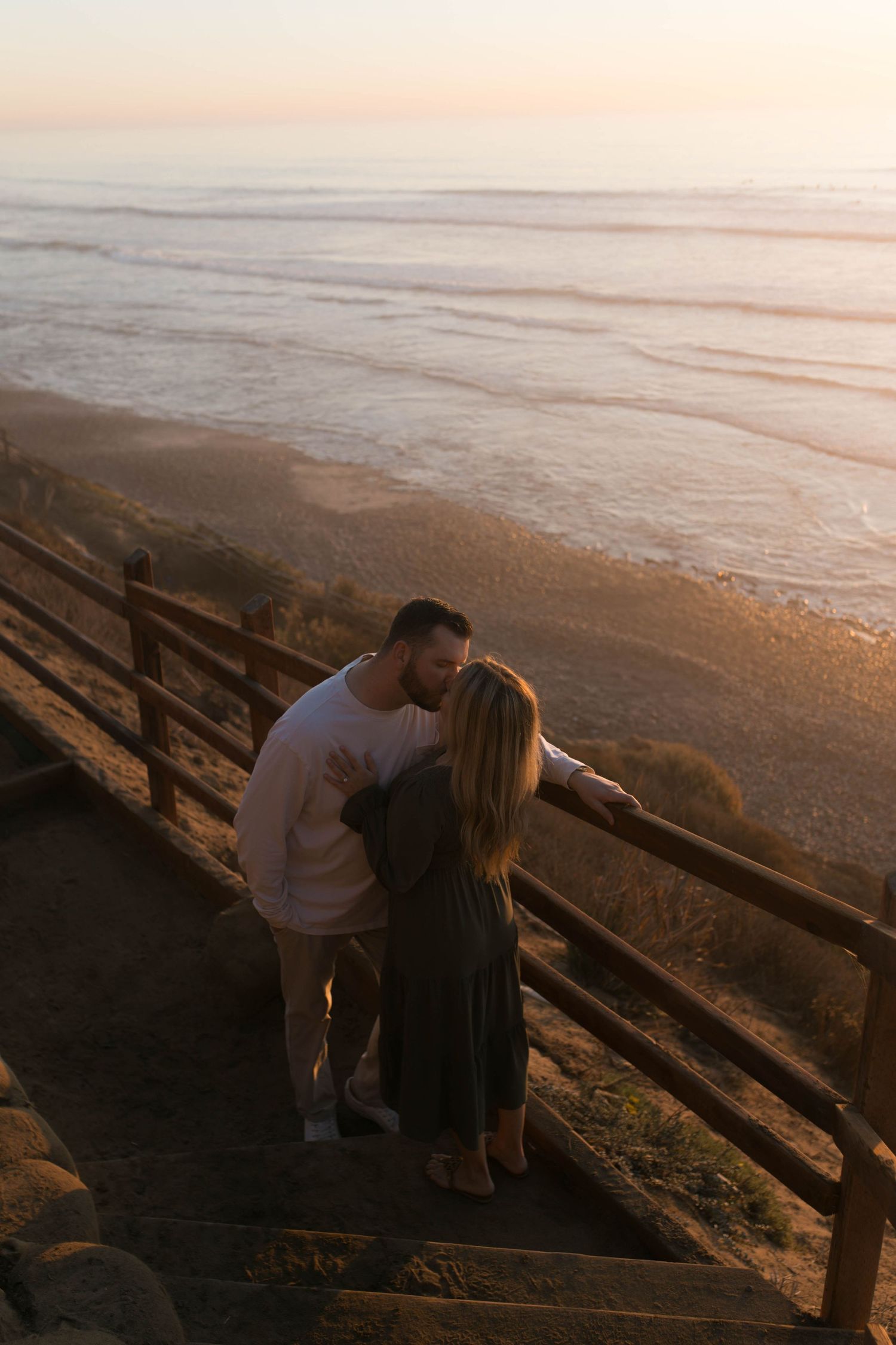 A couple shares an intimate moment at sunset on wooden stairs overlooking a misty beach.