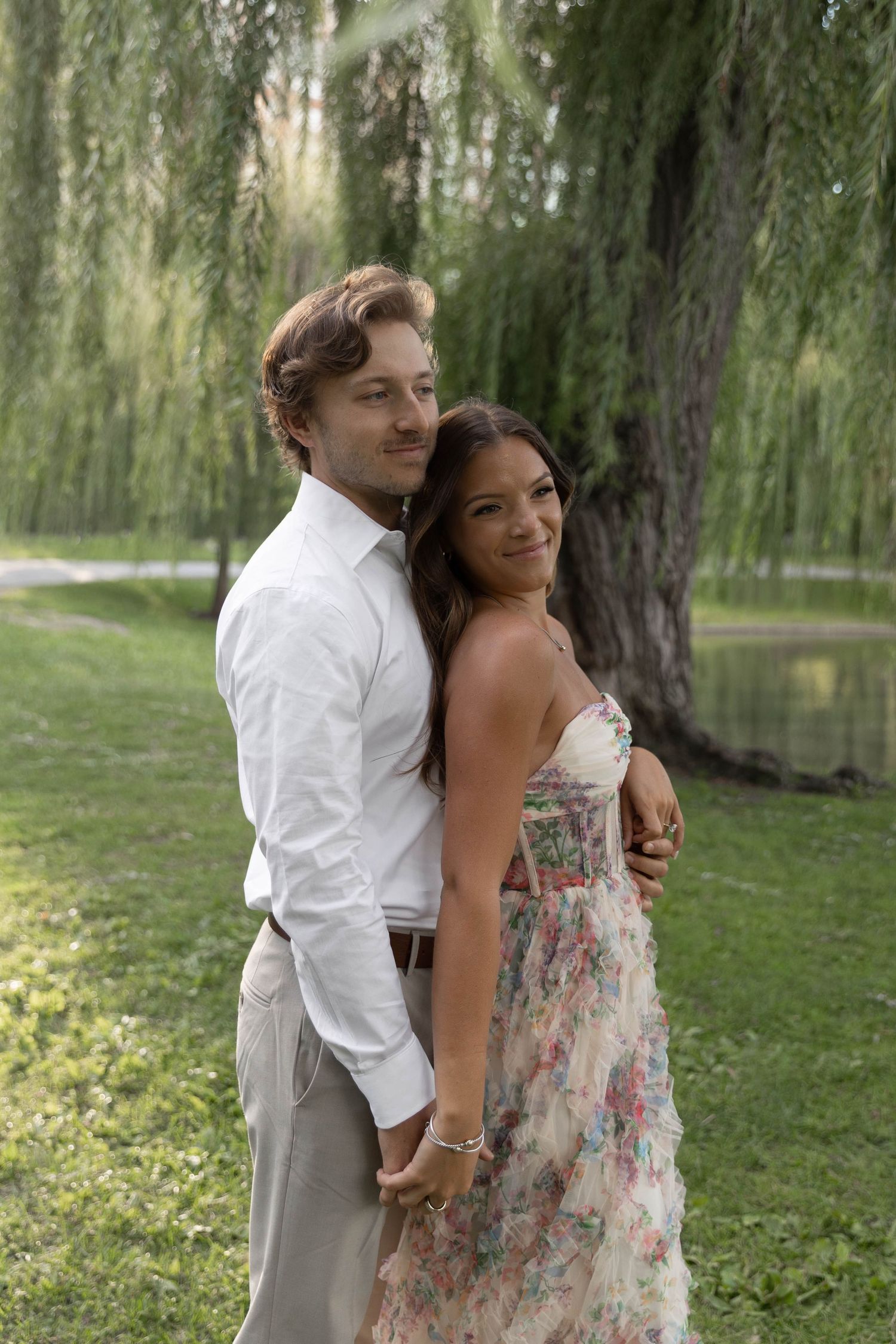 A romantic couple embraces under a weeping willow tree, with the woman wearing a floral dress and man in white shirt.