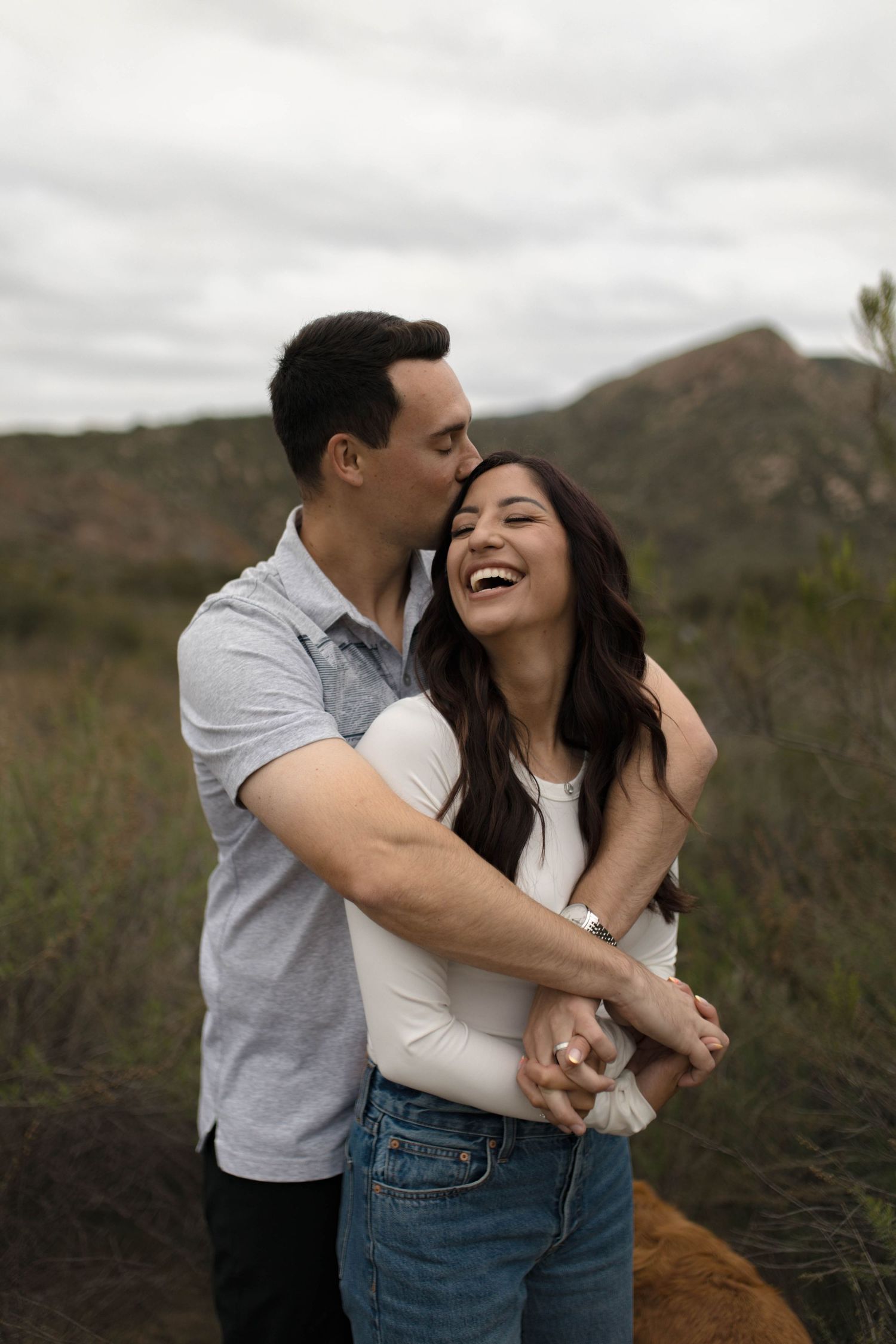 A joyful couple embraces and laughs together on a hiking trail with mountains in the background.