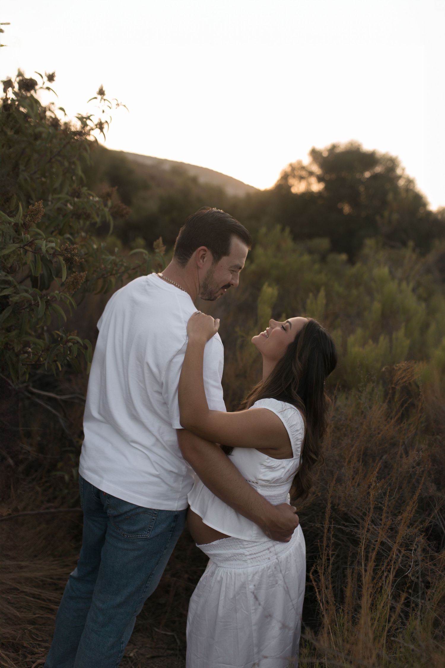 A romantic couple in white outfits embrace during sunset in a field of tall grass and wildflowers.