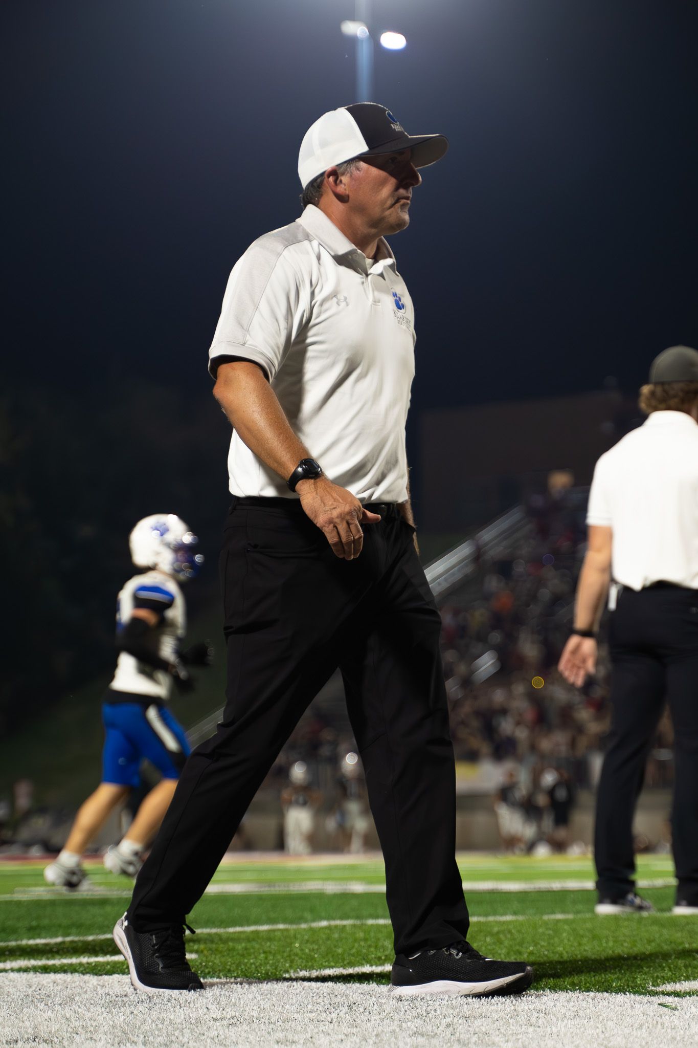 Coach in white polo shirt and black pants walks sideline during night football game.