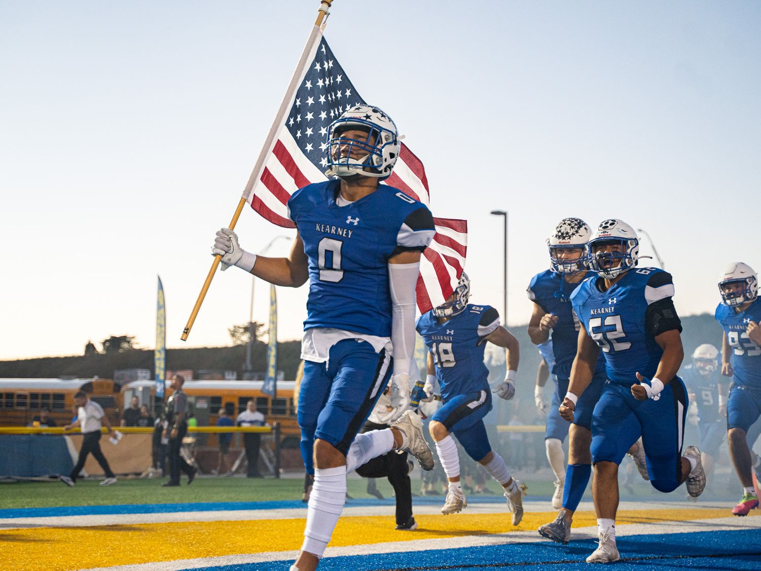Players run onto field carrying American flag before football game.