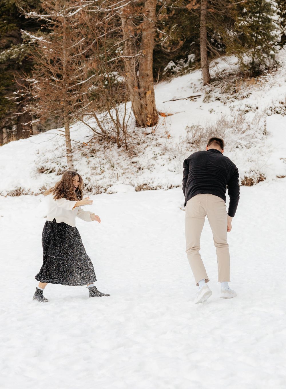 Playful couple having a snowball fight in a snowy winter landscape with pine trees.