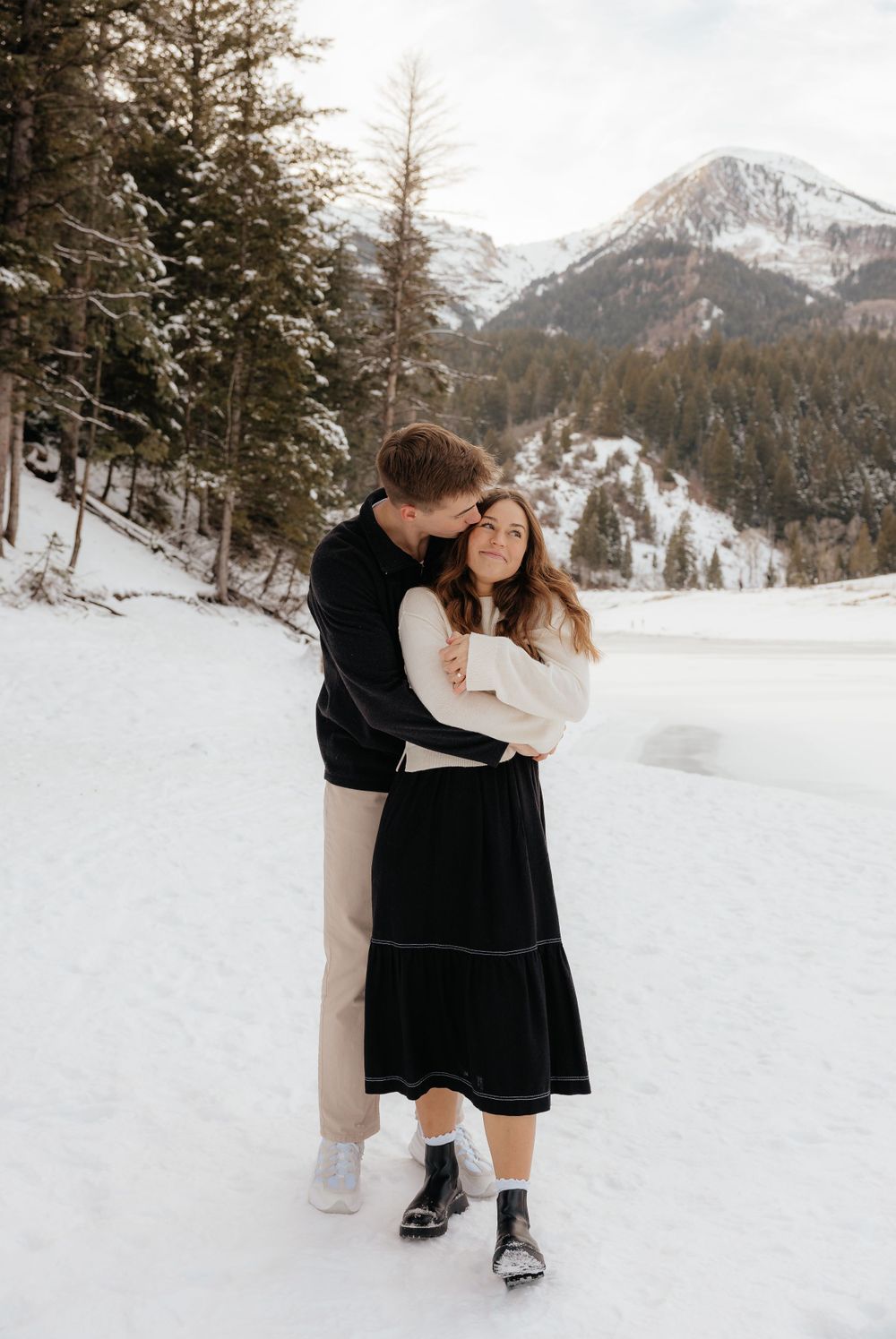 Man hugging woman from behind and kissing her head in a snowy mountain setting.