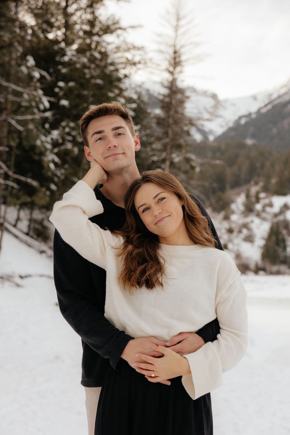 Couple posing together in a snowy mountain landscape, man embracing woman from behind.