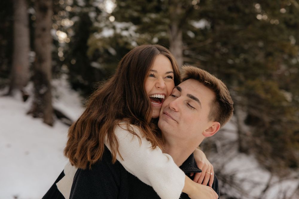 Joyful woman laughing while being carried piggyback by man in snowy winter forest setting.