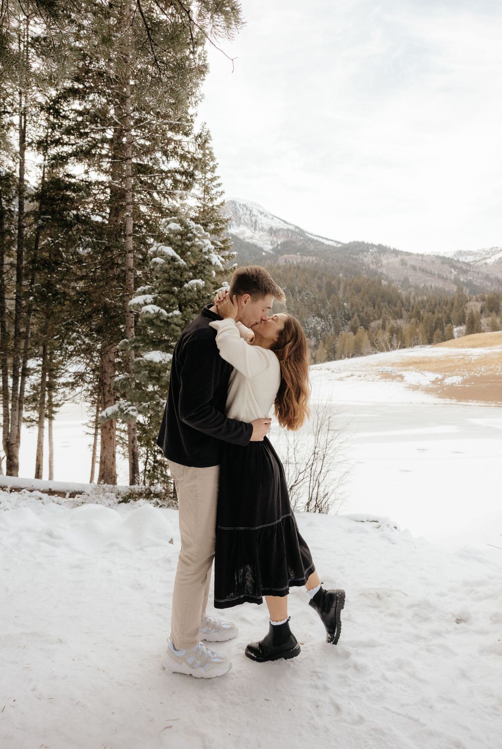 Couple sharing a kiss in snowy mountain landscape surrounded by pine trees and frozen lake.