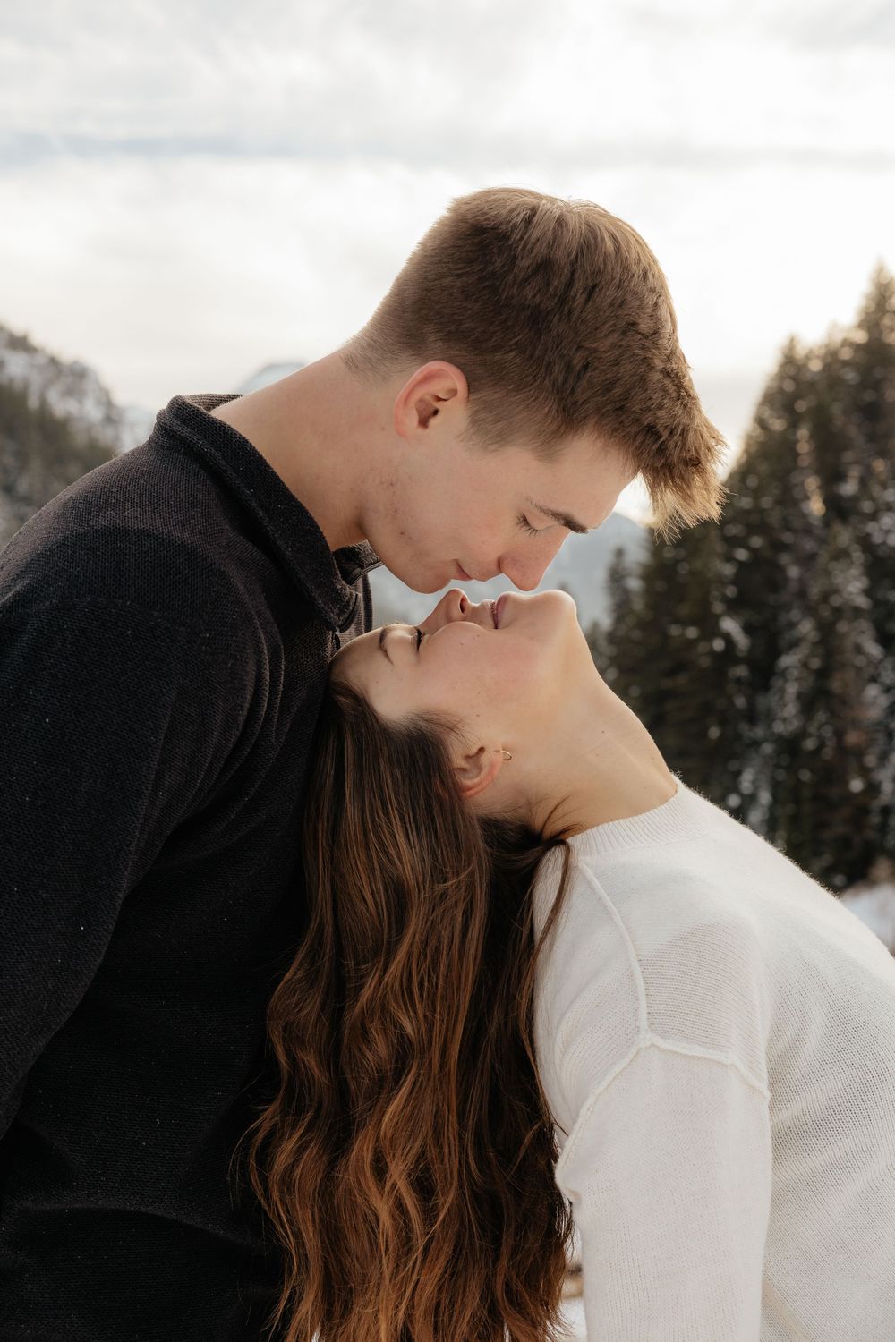 Romantic close-up of couple about to kiss with snowy pine forest and cloudy sky in background.
