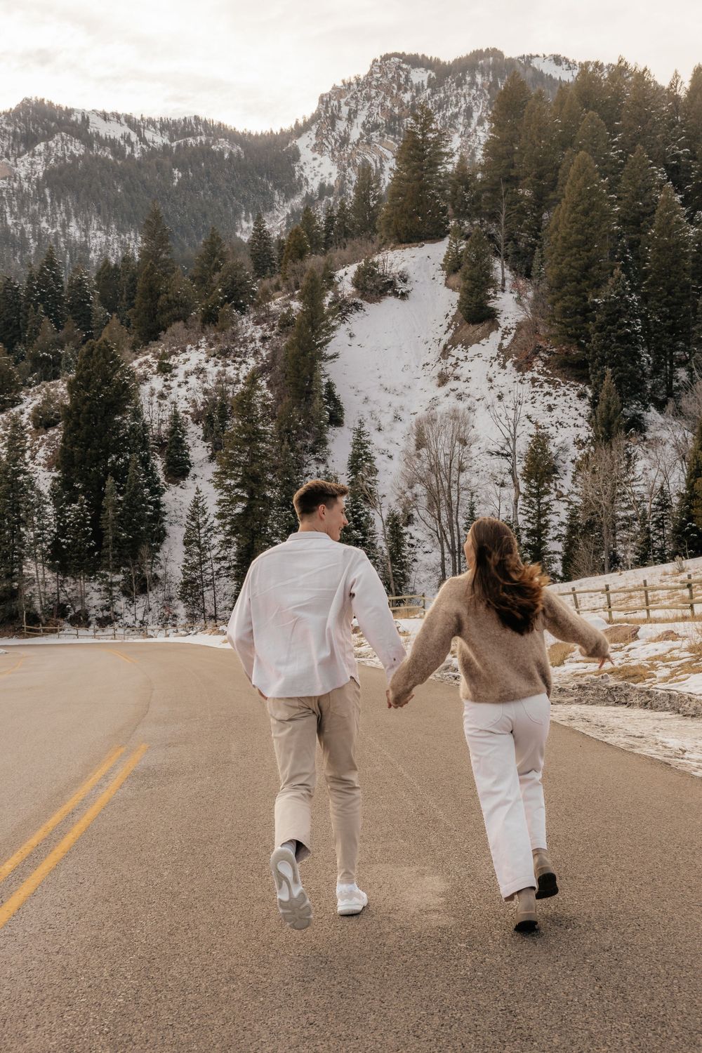 Couple walking hand in hand down mountain road in matching white outfits with snowy hillside behind.