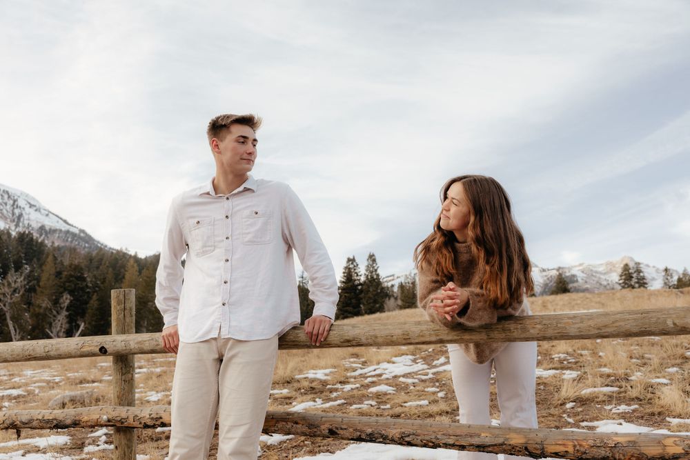 Young couple standing by a rustic wooden fence in a snowy mountain meadow on a cloudy day.