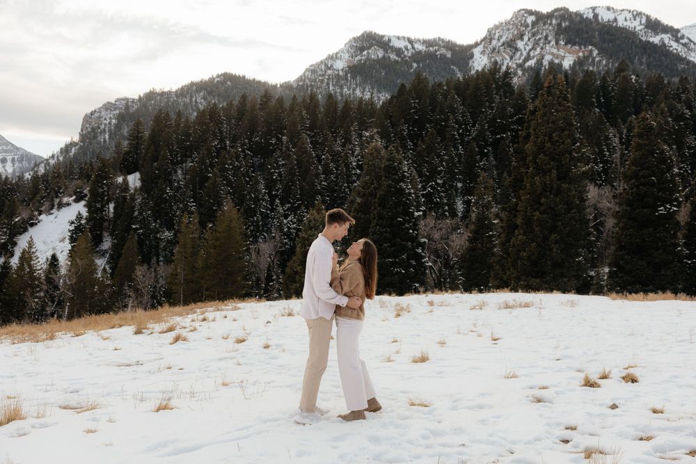 Engaged couple sharing a romantic kiss in a snowy mountain field surrounded by pine trees.