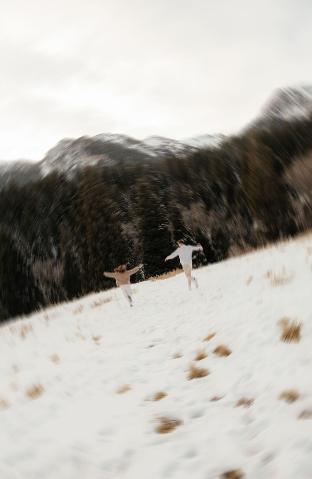 Couple running playfully up a snowy hillside with mountains and pine trees in background.