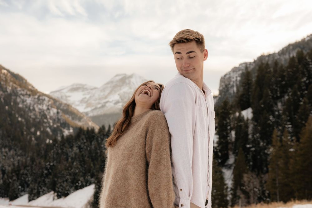Woman leaning on mans shoulder laughing together in a snowy mountain landscape with pine trees.