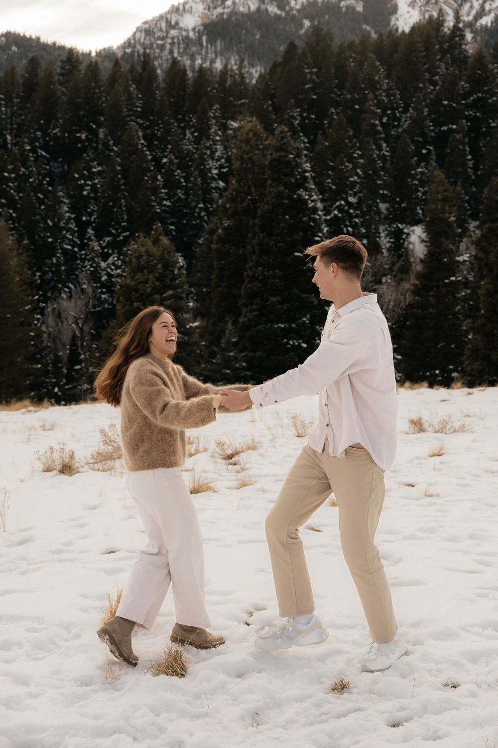 Happy couple dancing and spinning together in a snowy mountain field with evergreen trees.