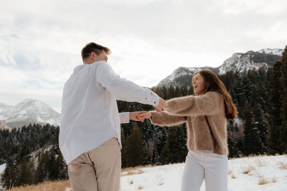 Couple playfully holding hands and spinning in a snowy mountain meadow with pine trees.