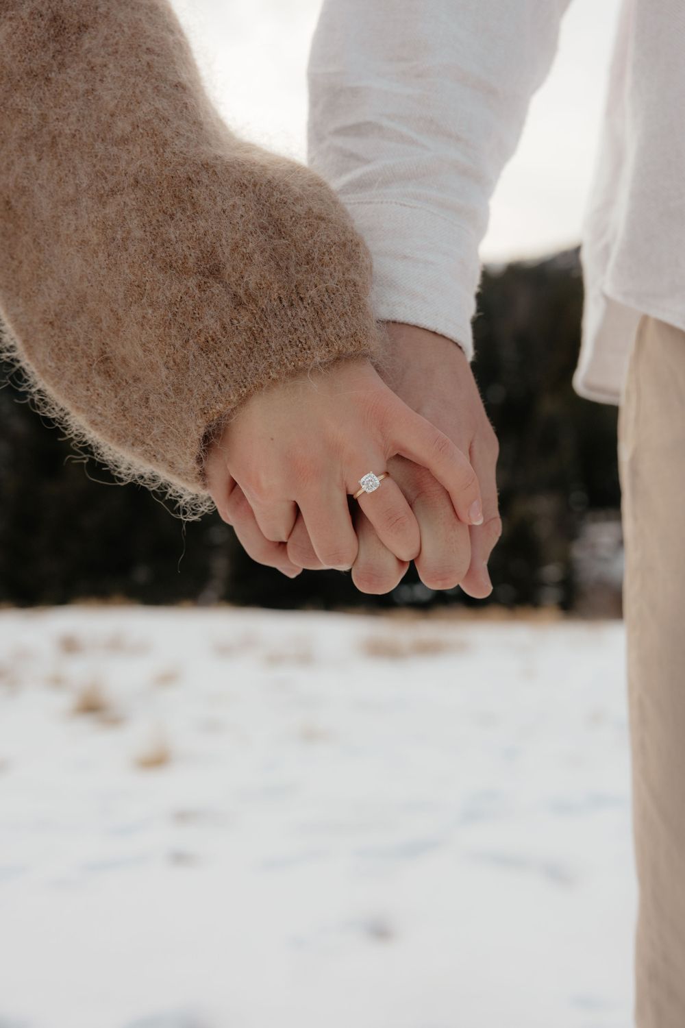Close-up of engaged couples hands showing diamond engagement ring in a snowy winter setting.
