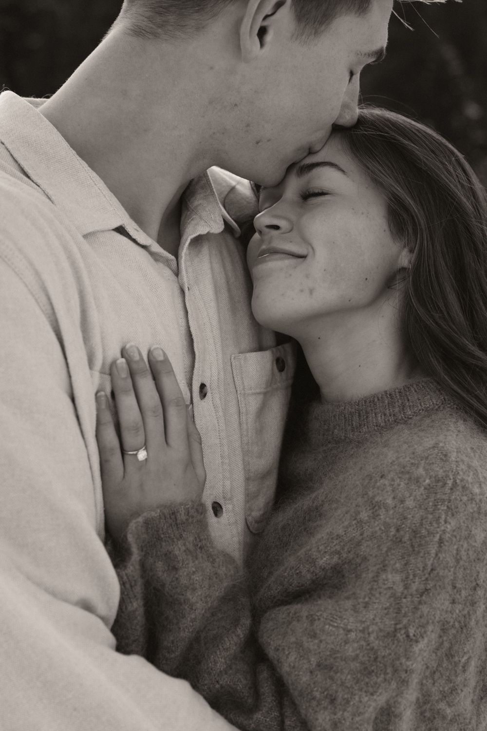 Black and white photo of a couple embracing, woman smiling with engagement ring visible on hand.