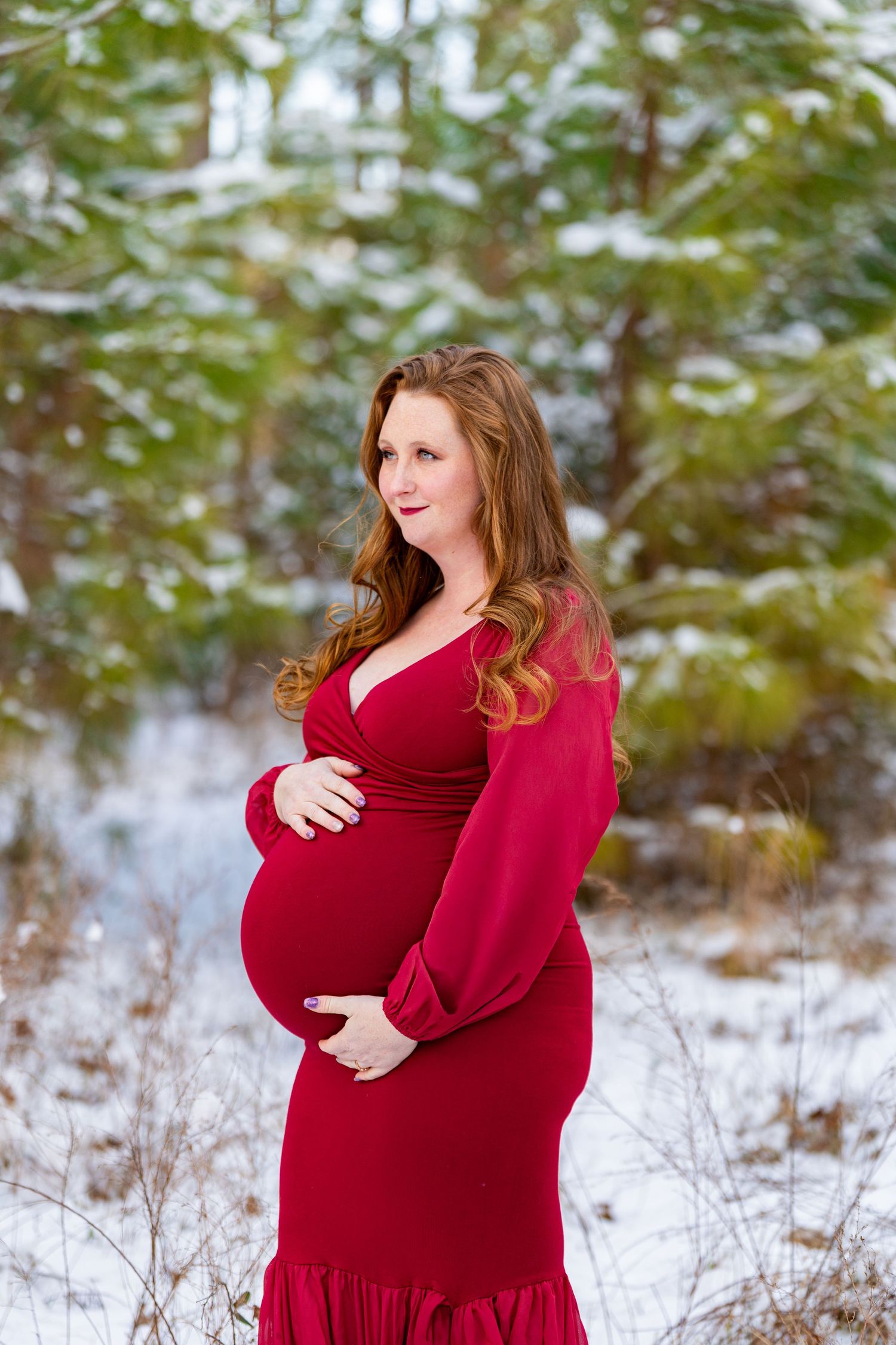 A person in a flowing red maternity dress poses outdoors in a snowy winter setting with evergreen trees in background.