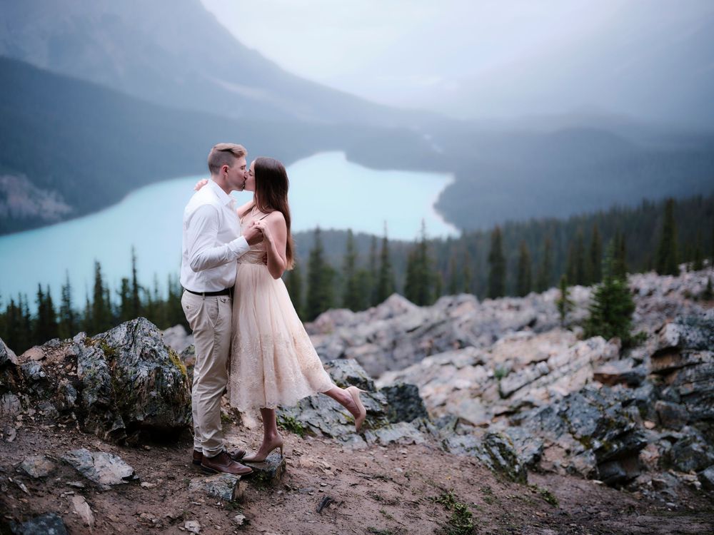 A couple shares a romantic moment on a mountain overlook above a turquoise glacial lake surrounded by evergreen forest in the Rockies