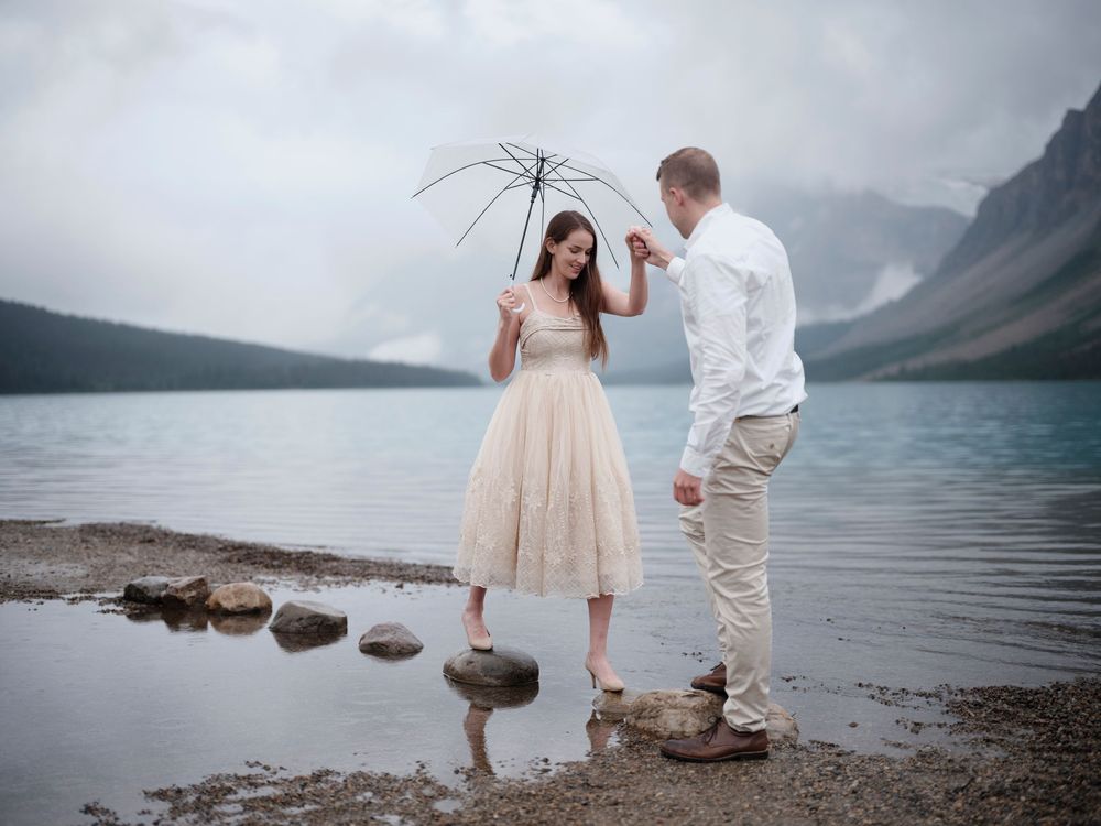Romantic couple stands with umbrella by misty mountain lake on rocky shoreline in vintage style dress and attire engagement photos Banff