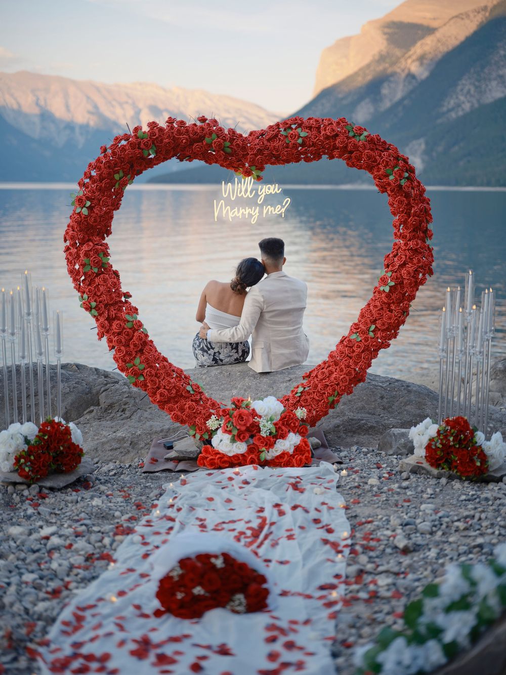 Red rose heart-shaped arch frames a romantic proposal scene against a scenic mountain lake backdrop in Banff