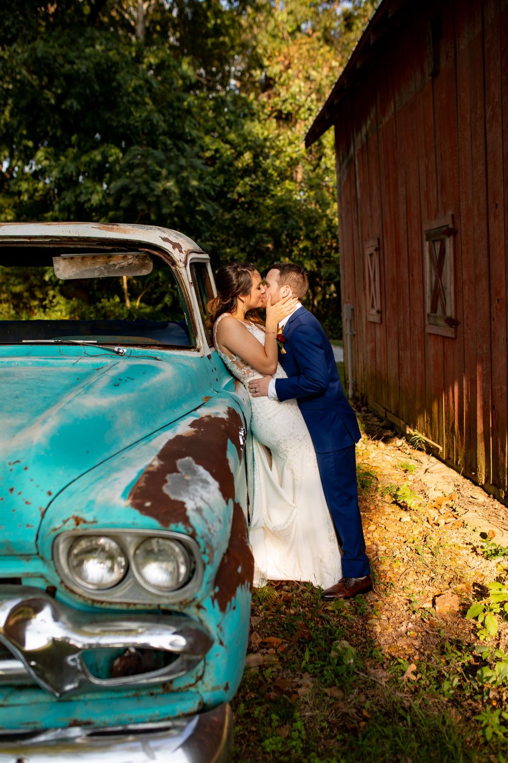 Couple shares romantic moment beside vintage turquoise truck near rustic red barn in autumn setting.