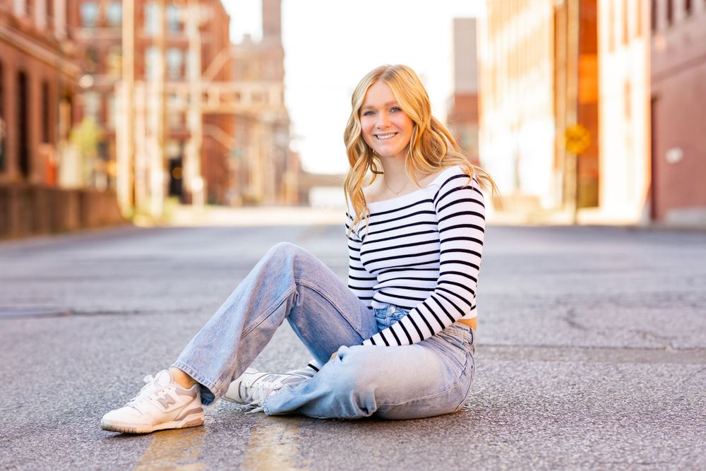 A person in a striped shirt and jeans sits casually on a city street during golden hour.